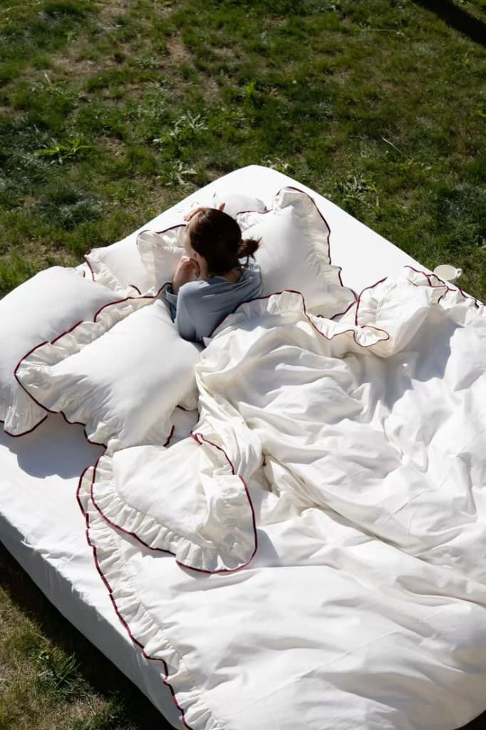 A person resting on white ruffled bedding with red trim, outdoors on a sunny green lawn.